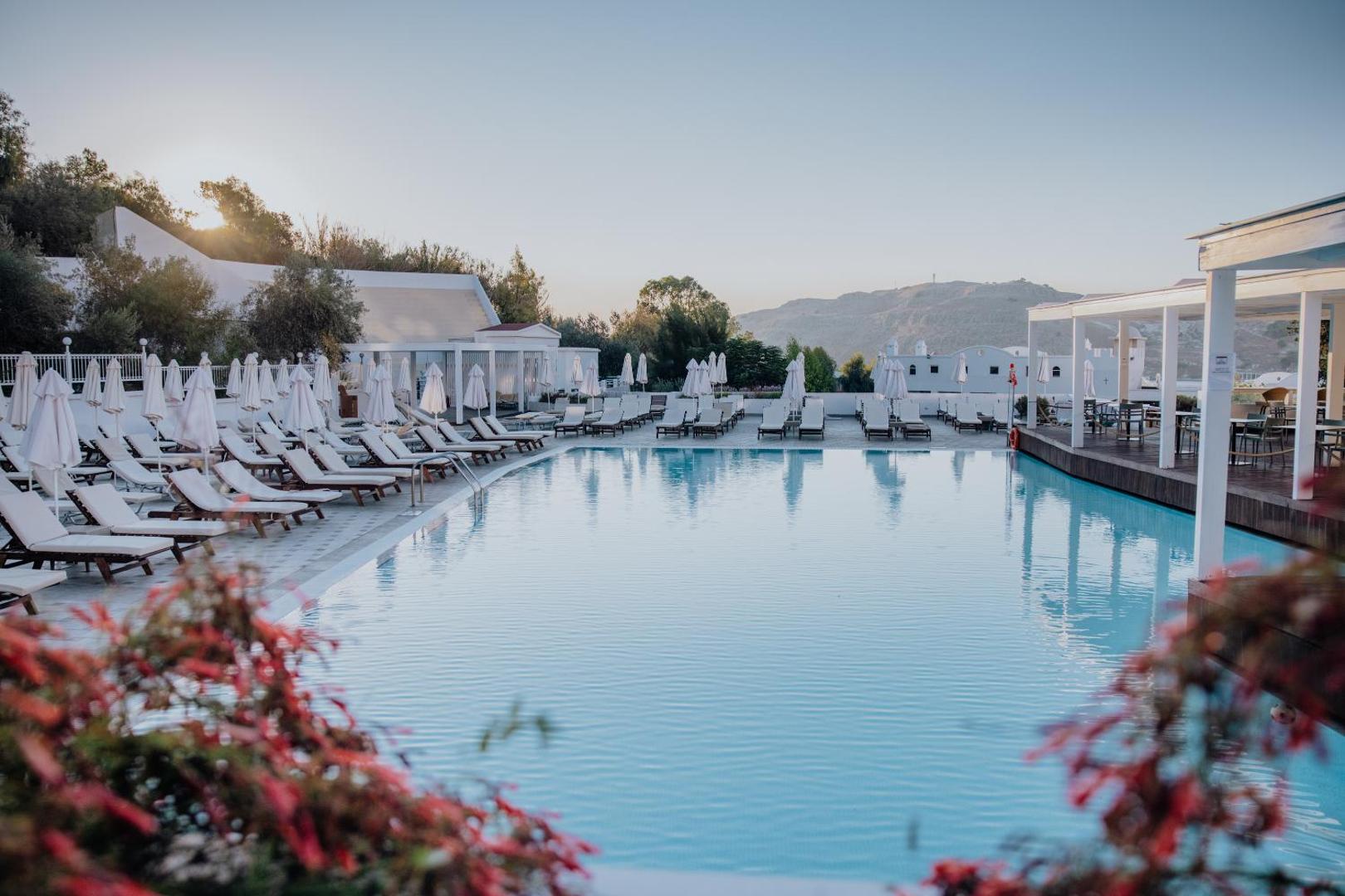 Lindos Village Resort & Spa galvenais āra baseins saullēkta laikā Rodas salā ar baltiem saulessargiem un kalnu panorāmu fonā