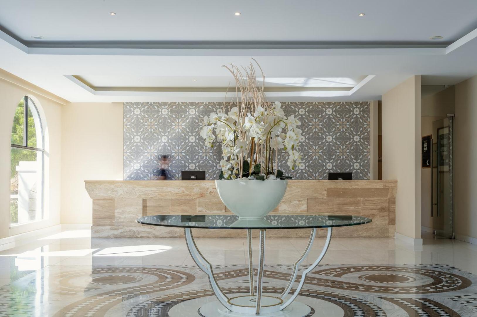 Lindos Village Resort & Spa gaišā recepcijas zona ar marmora apdari, ziedu kompozīciju un rakstainu flīžu sienu Rodas salā