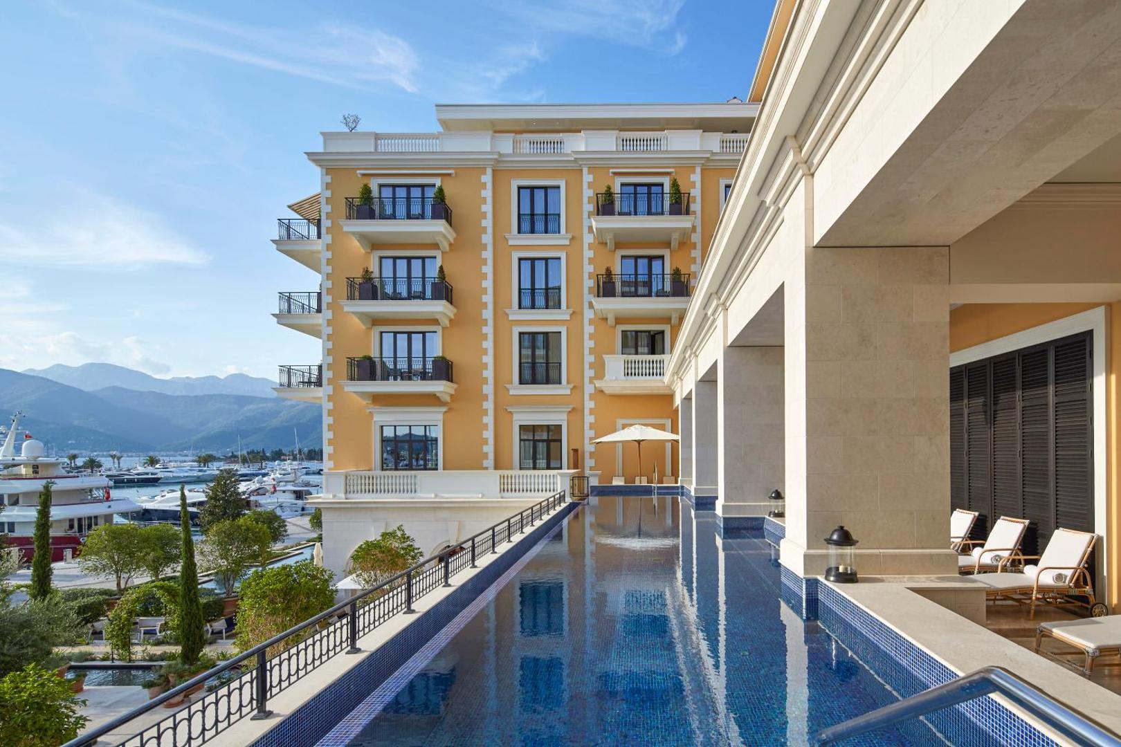 Regent Porto Montenegro viesnīcas fasāde un baseins piekrastē ar skatu uz jahtu piestātni un kalniem saulainā dienā