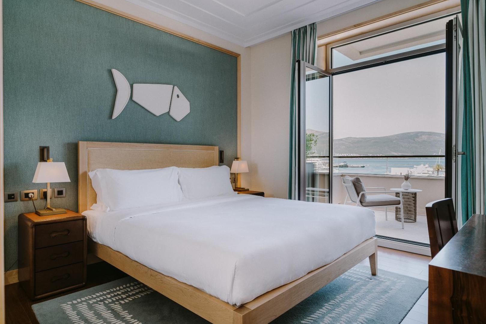 Regent Porto Montenegro numurs ar skatu uz jūru un balkonu — gaišs interjers ar zilu akcenta sienu un koka mēbelēm