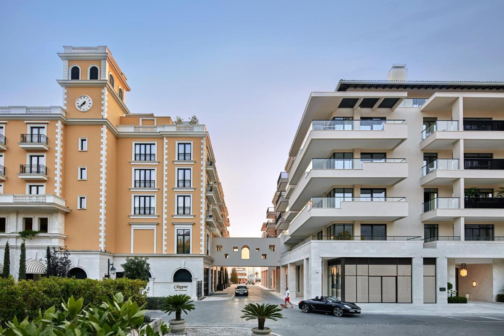 Regent Porto Montenegro viesnīcas fasāde ar pulksteņa torni un modernu piebūvi — ieeja kūrortā ar palmām un luksusa automašīnām