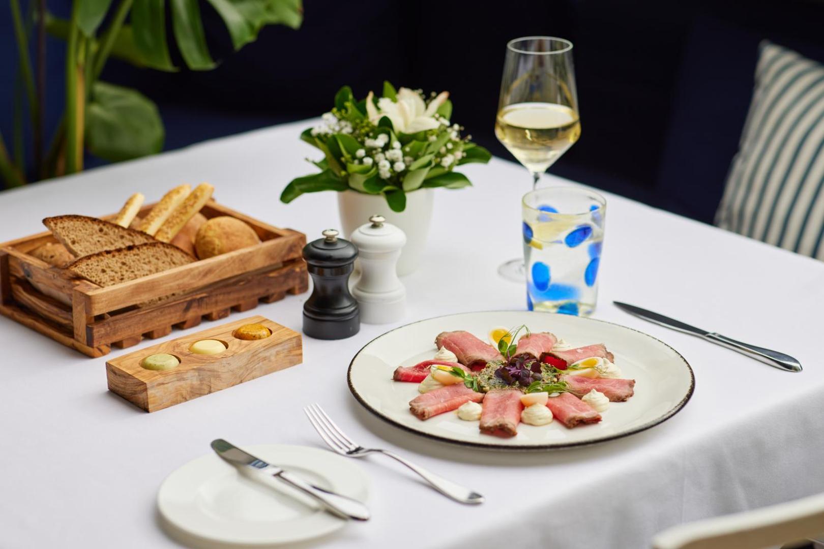 Gourmet uzkoda uz balta galdauta Regent Porto Montenegro restorānā — gaļas karpačo ar mērcēm, vīna glāzi un svaigu maizi