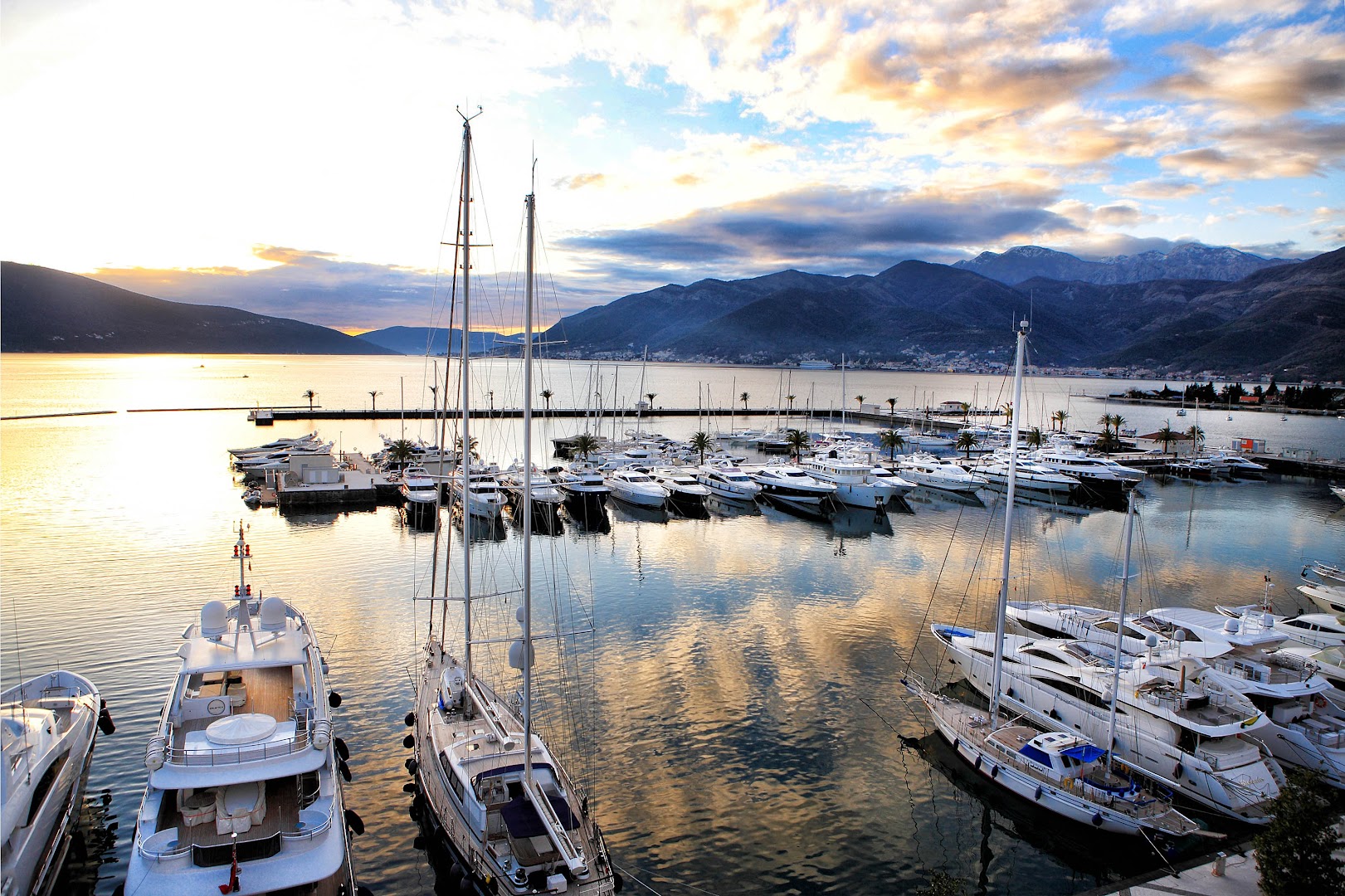 Regent Porto Montenegro skats uz jahtu piestātni saulrietā Tivatas līcī, Melnkalnē — greznas jahtas un kalnu panorāma vakara gaismā