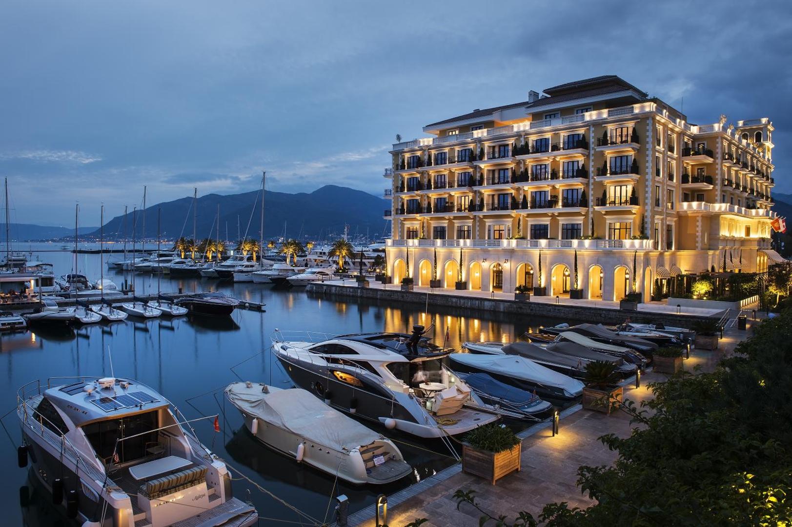 Regent Porto Montenegro izgaismota fasāde vakarā pie jahtu piestātnes — luksusa jahtas pietauvotas tieši pie viesnīcas restorānu terasēm