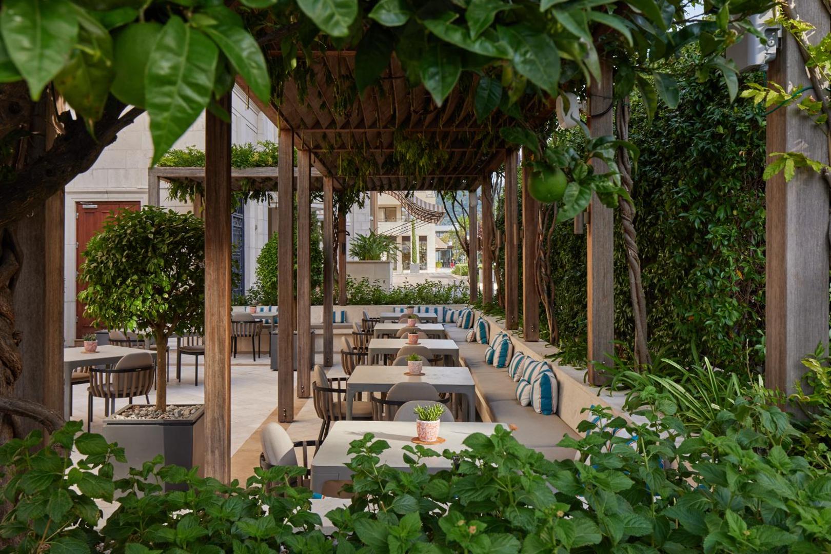 Regent Porto Montenegro dārza terase zem zaļas lapotnes ar koka konstrukcijām un ērtiem sēdekļiem atpūtai brīvā dabā