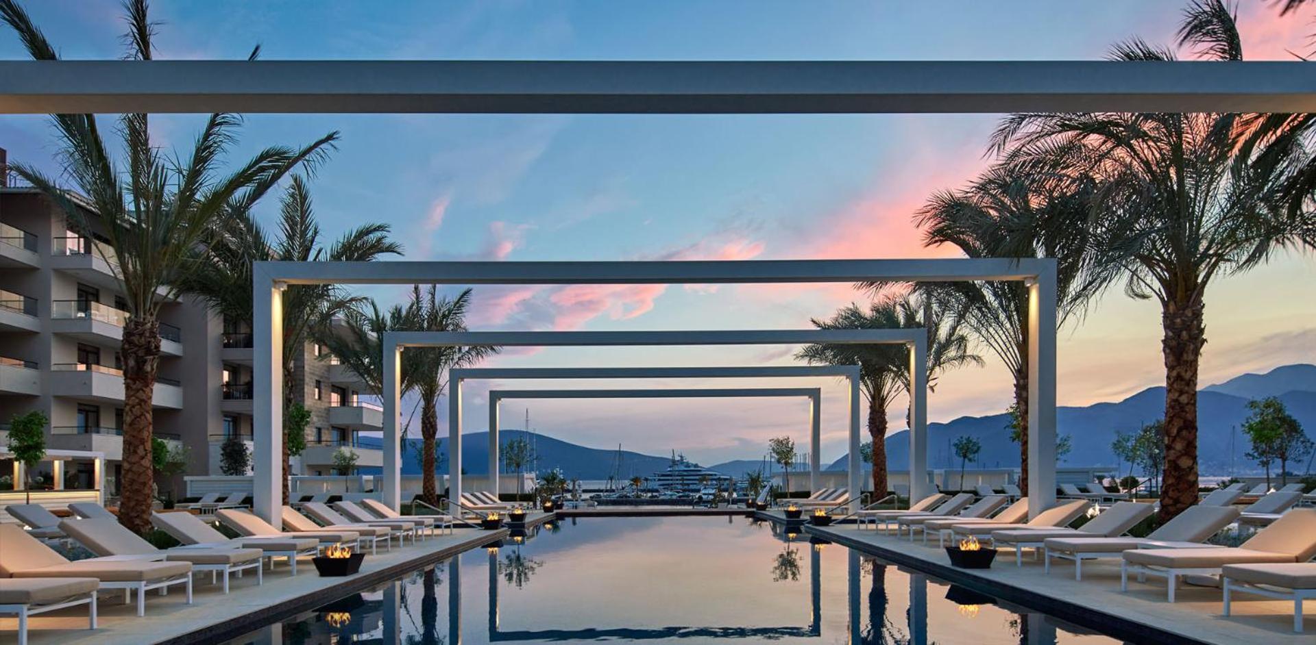 Regent Porto Montenegro baseina zona vakarā — panorāmas skats uz jahtām un kalniem zem rozā saulrieta debesīm