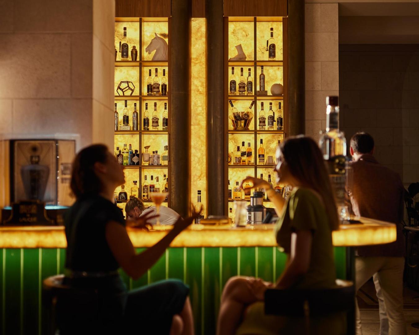 Regent Porto Montenegro viesnīcas bārs ar izgaismotu dzērienu sienu un viesiem pie letes — eleganta vakara atmosfēra Melnkalnē