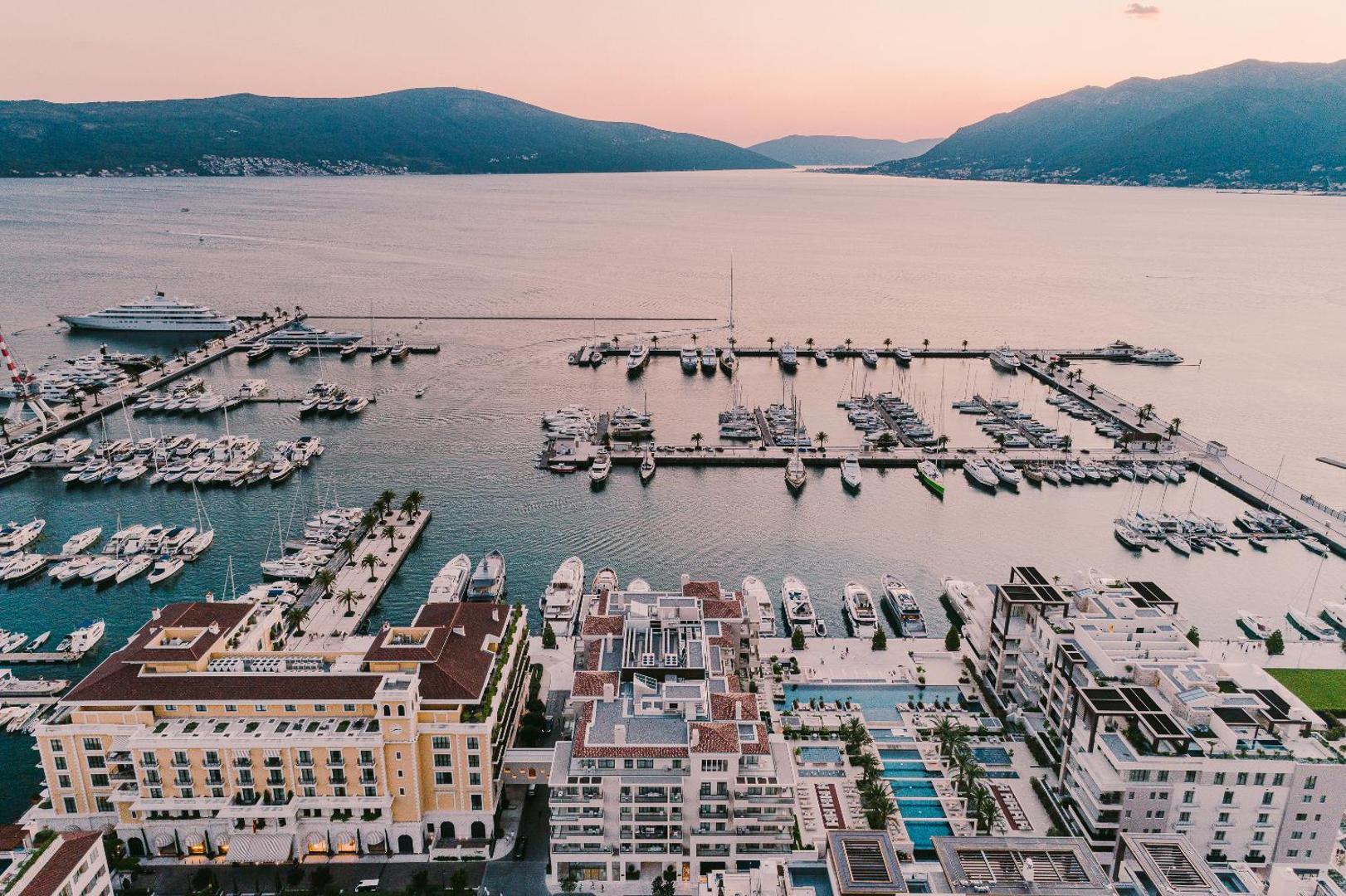 Regent Porto Montenegro panorāmas skats uz jahtu ostu un āra baseiniem saulrietā — kalnu ainava fonā pie Tivatas līča