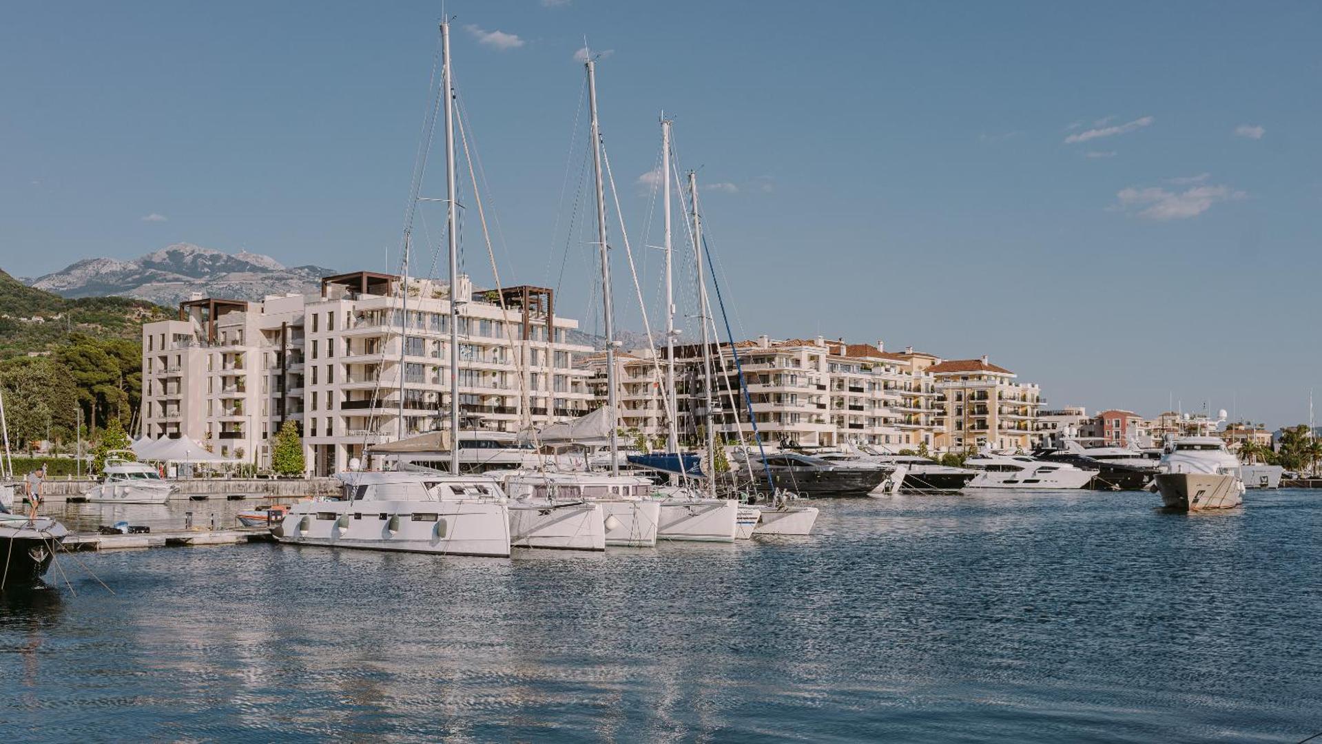 Baltas jahtas un katamarāni Regent Porto Montenegro piestātnē uz viesnīcas ēku un kalnu fona saulainā dienā