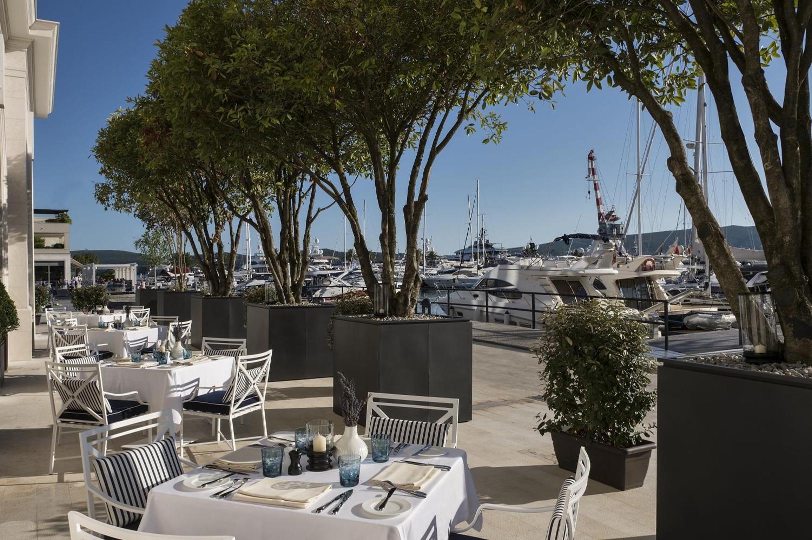 Regent Porto Montenegro āra terase ar galdiņiem pie jahtu piestātnes — koku ēna un skats uz ostu saulainā dienā