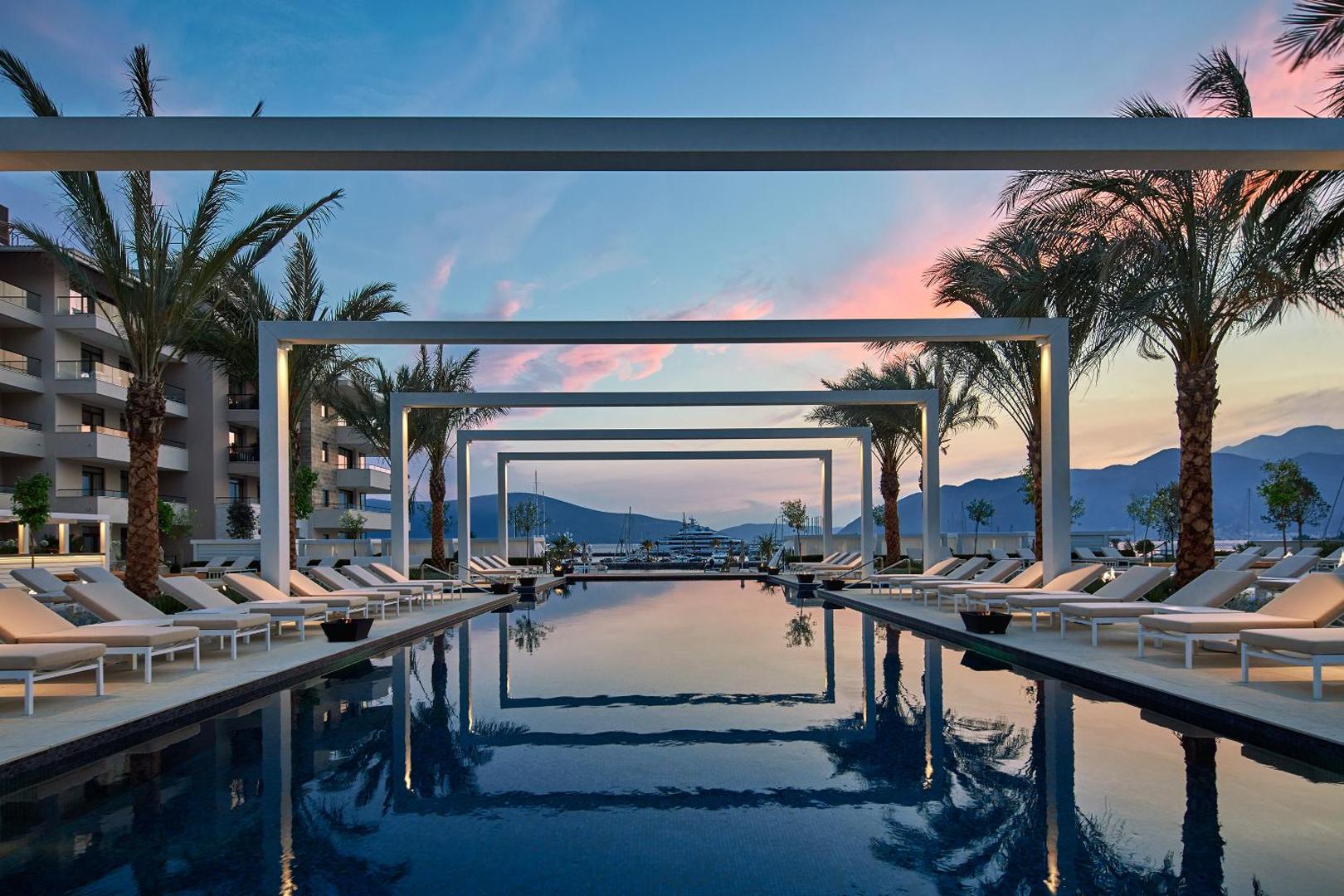 Regent Porto Montenegro āra baseins saulrietā ar skatu uz jahtu piestātni un kalniem — eleganti sauļošanās krēsli un palmas