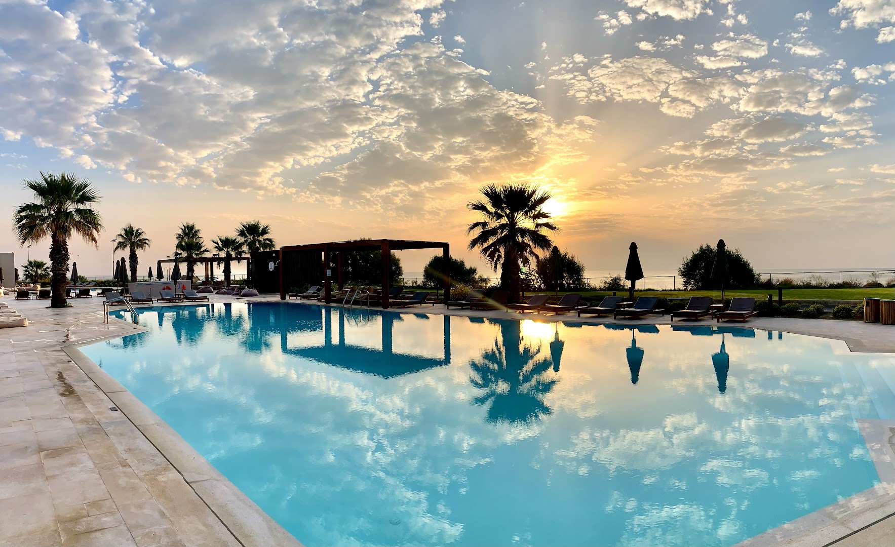 Saulriets pie Elissa Lifestyle Resort baseina — debesu atspulgs ūdenī, palmas un vakara atmosfēra Rodas piekrastē
