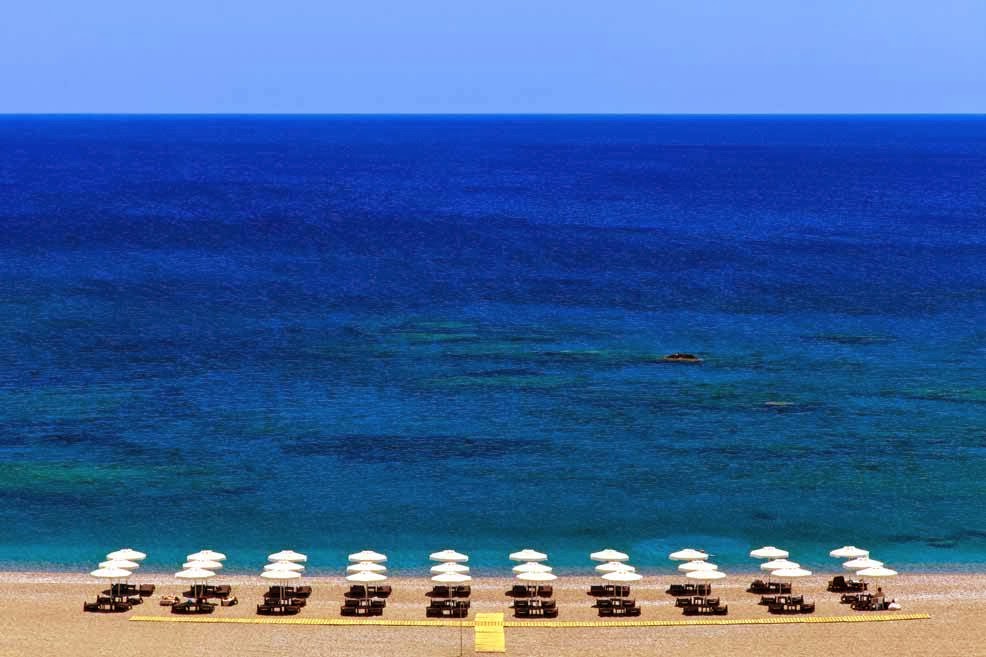 Boutique 5 Hotel & Spa privātā pludmale Rodas salā ar baltiem saulessargiem un sauļošanās krēsliem pie dzidri zilā Vidusjūras ūdens