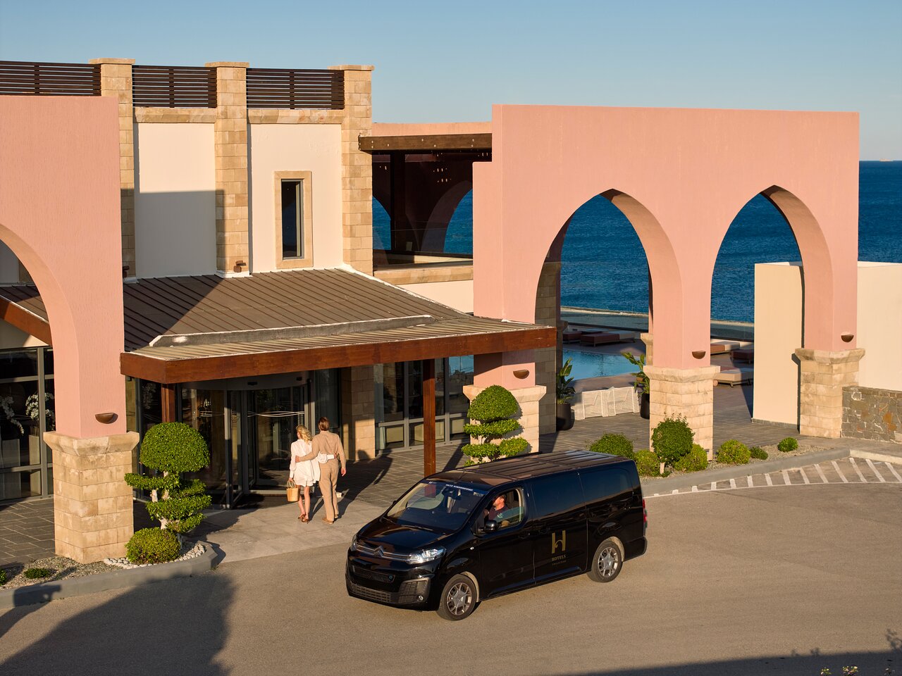 Viesu ierašanās Boutique 5 Hotel & Spa Rodā — viesnīcas ieeja ar rozā arkām, transfēra auto un skatu uz jūru