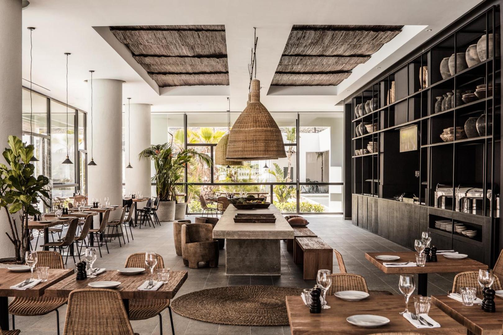 Casa Cook Rhodes boutique viesnīcas restorāna interjers ar dabīgā koka mēbelēm, pītiem gaismekļiem un lieliem logiem uz dārzu