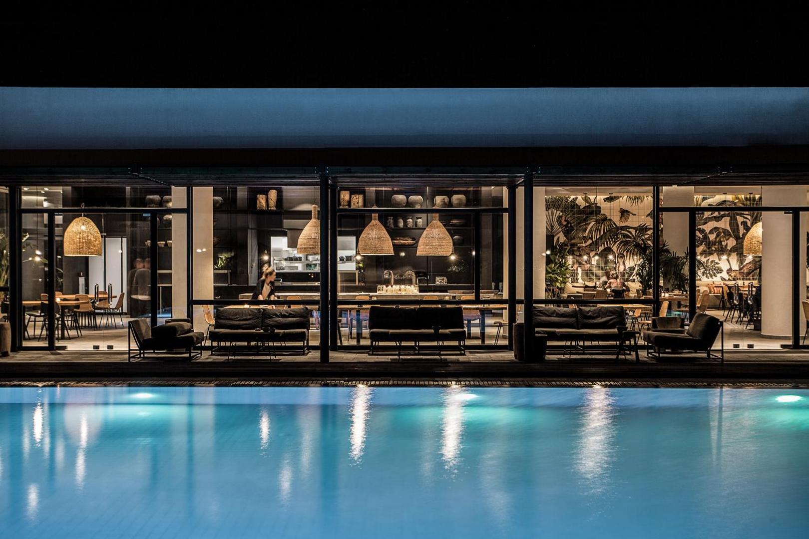 Casa Cook Rhodes restorāna un lobby zona vakarā ar apgaismotu baseinu, modernu interjeru un dizaina lampām Rodas salā.