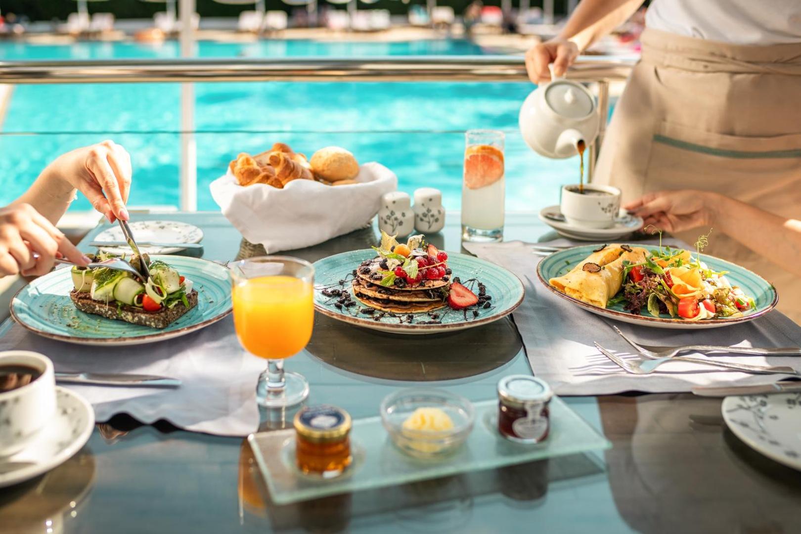 Brokastis uz terases Mitsis Faliraki Beach Hotel & Spa ar skatu uz baseinu — pankūkas, avokado maize un svaiga sula