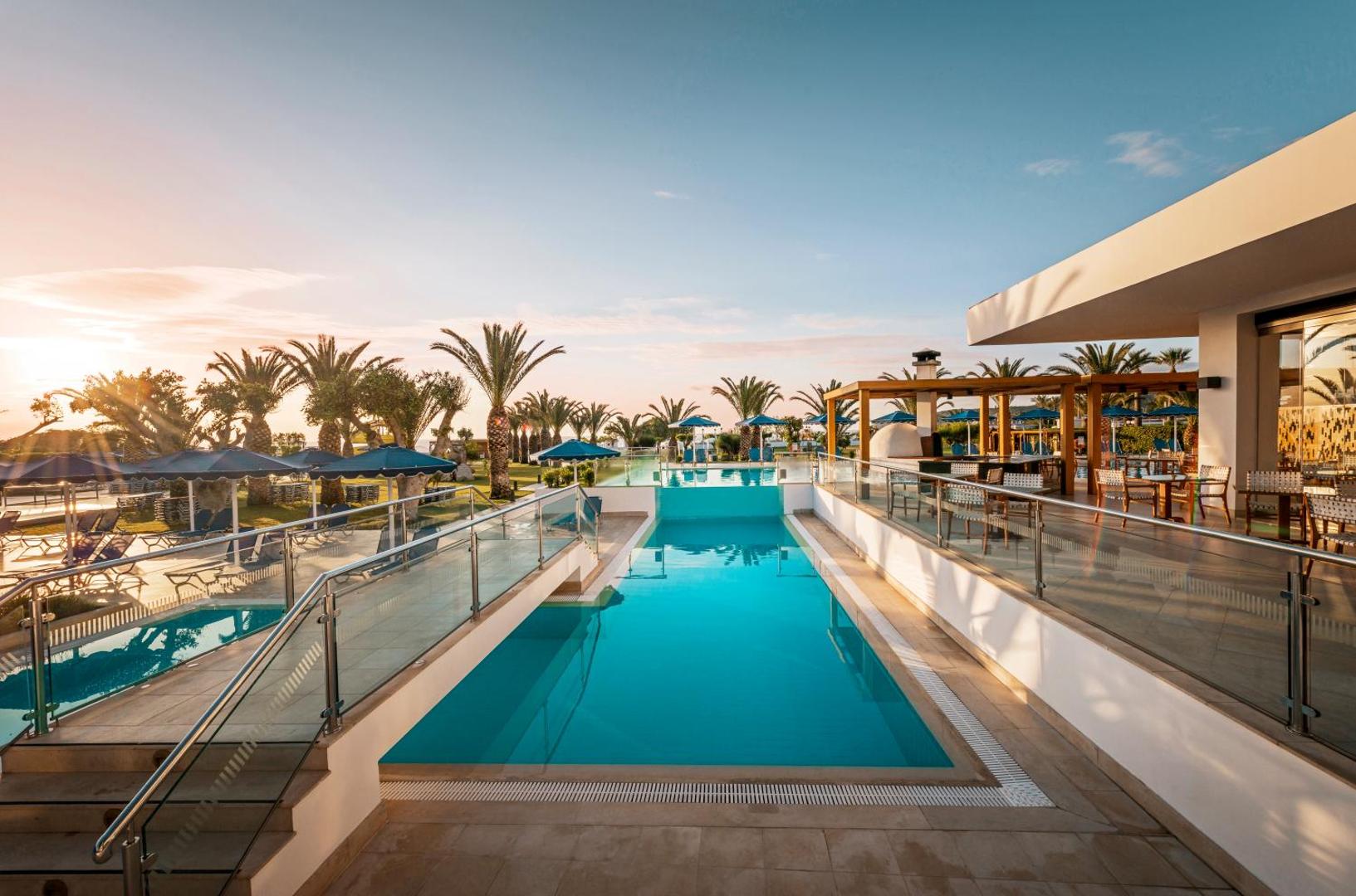 Mitsis Faliraki Beach Hotel & Spa baseins ar stikla norobežojumu un terasi Rodas salā saulrieta laikā — skats uz dārzu un palmām