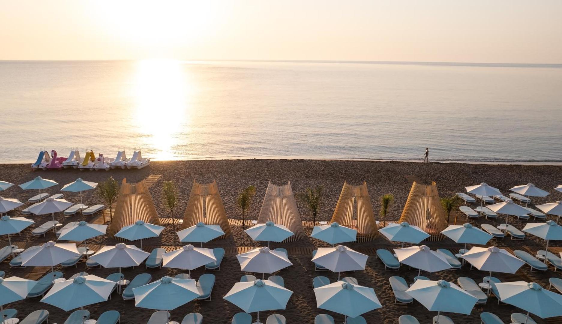 Mitsis Faliraki Beach Hotel & Spa privātā pludmale Rodas salā saullēkta laikā ar gaiši ziliem saulessargiem un koka atpūtas teltīm