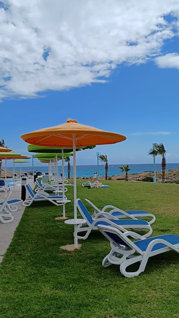 Aktea Beach Village atpūtas zona ar ziliem sauļošanās krēsliem un krāsainiem saulessargiem uz zaļas zāles piekrastē, Kiprā.