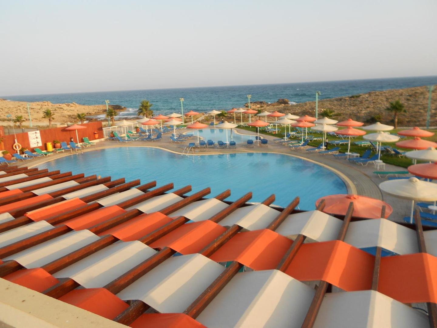 Skats no augšas uz Aktea Beach Village baseinu kompleksu un piekrasti — dekoratīvas nojumes un daudz saulessargu atpūtas zonā