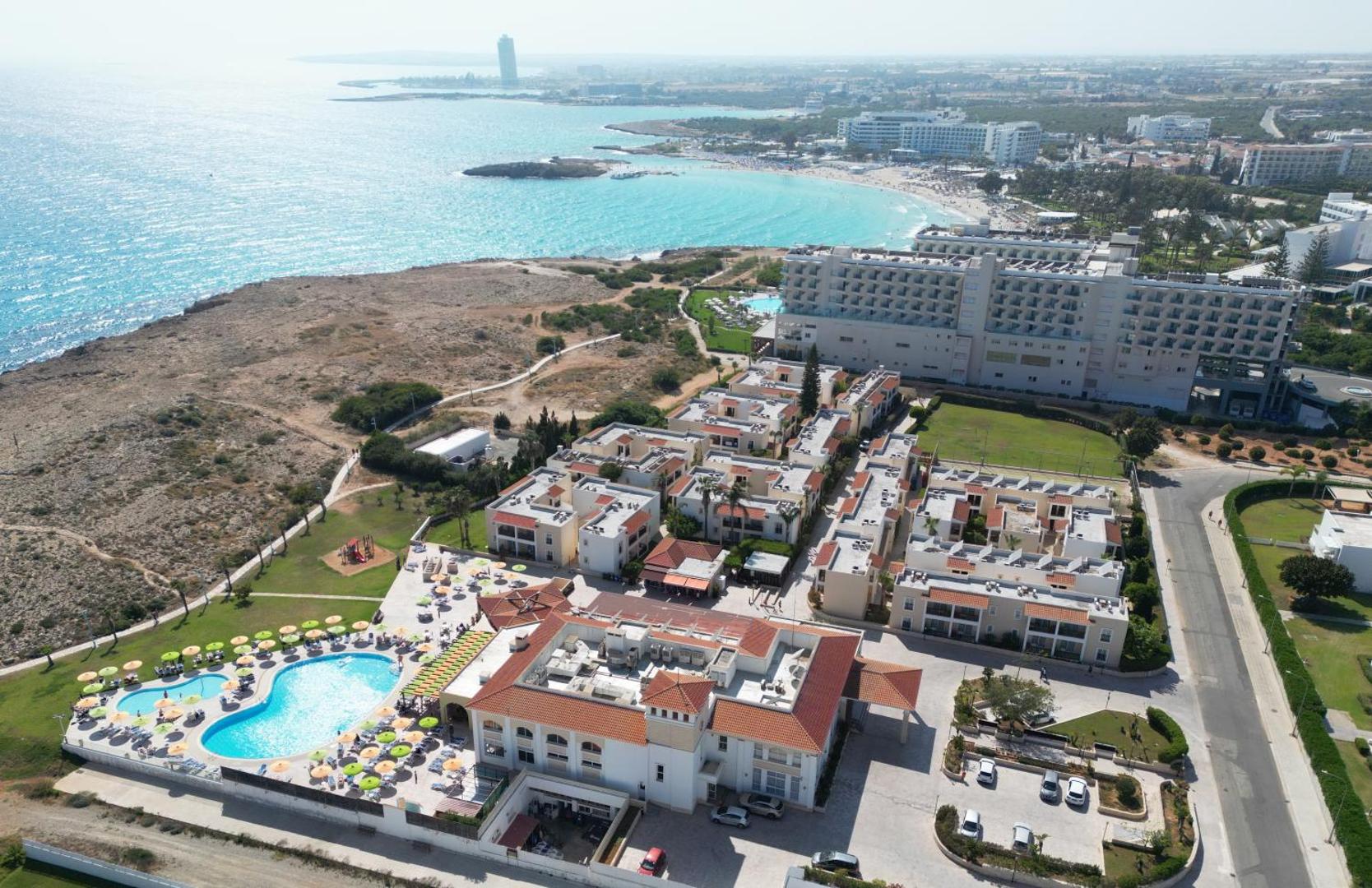 Aktea Beach Village teritorijas skats no augšas — redzams baseins ar saulessargiem, viesnīcas korpusi un tuvējā smilšu pludmale līča krastā
