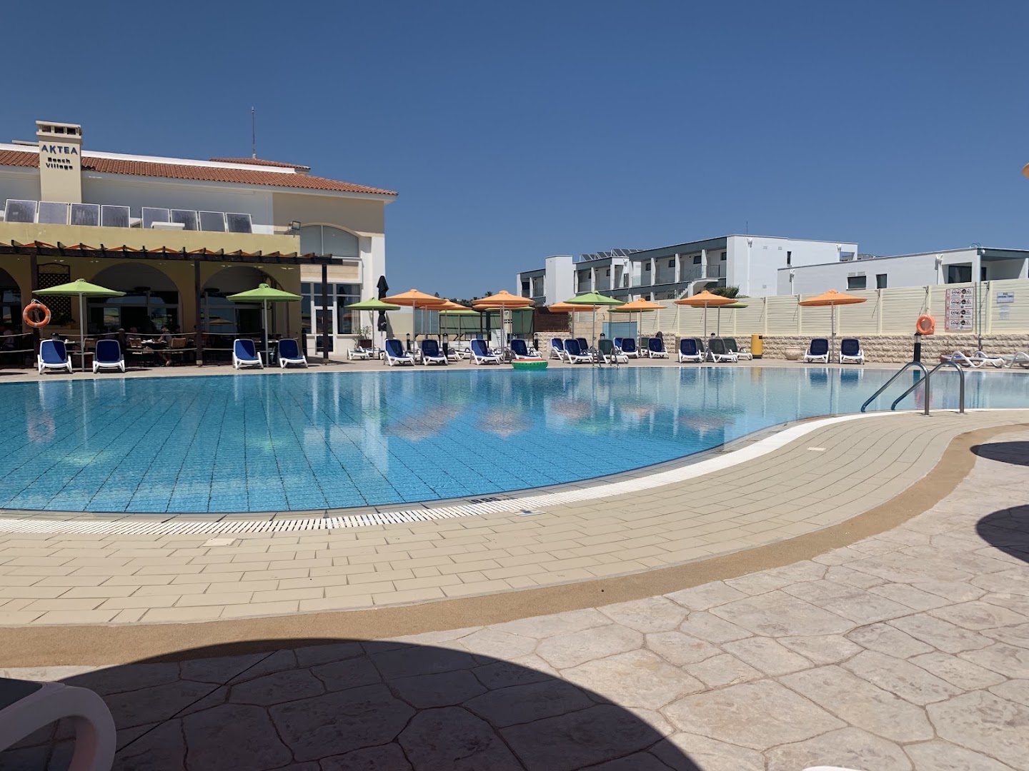 Aktea Beach Village galvenais baseins pie viesnīcas ēkas — saulaina terase ar krāsainiem saulessargiem un ieeju restorānā