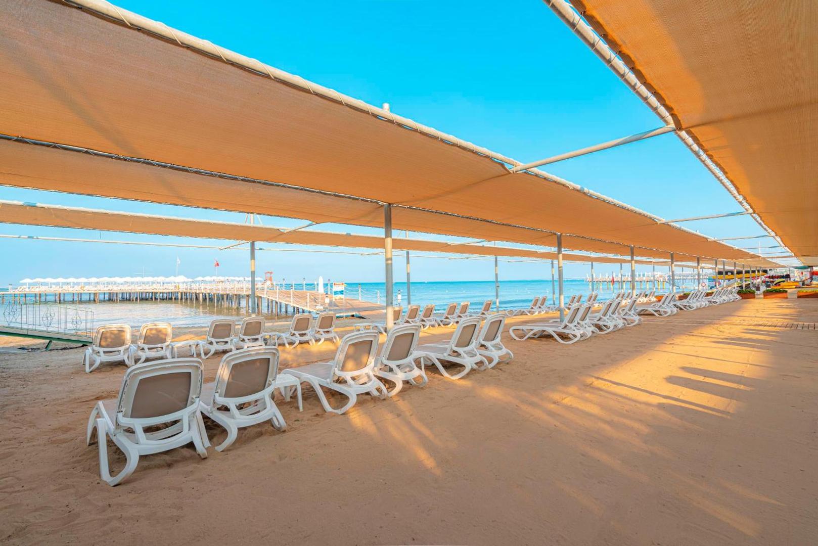 Kirman Sidemarin Beach & Spa smilšu pludmales zona ar plašu tentu nojumi un sauļošanās krēsliem Sidē — skats uz jūru un laipu