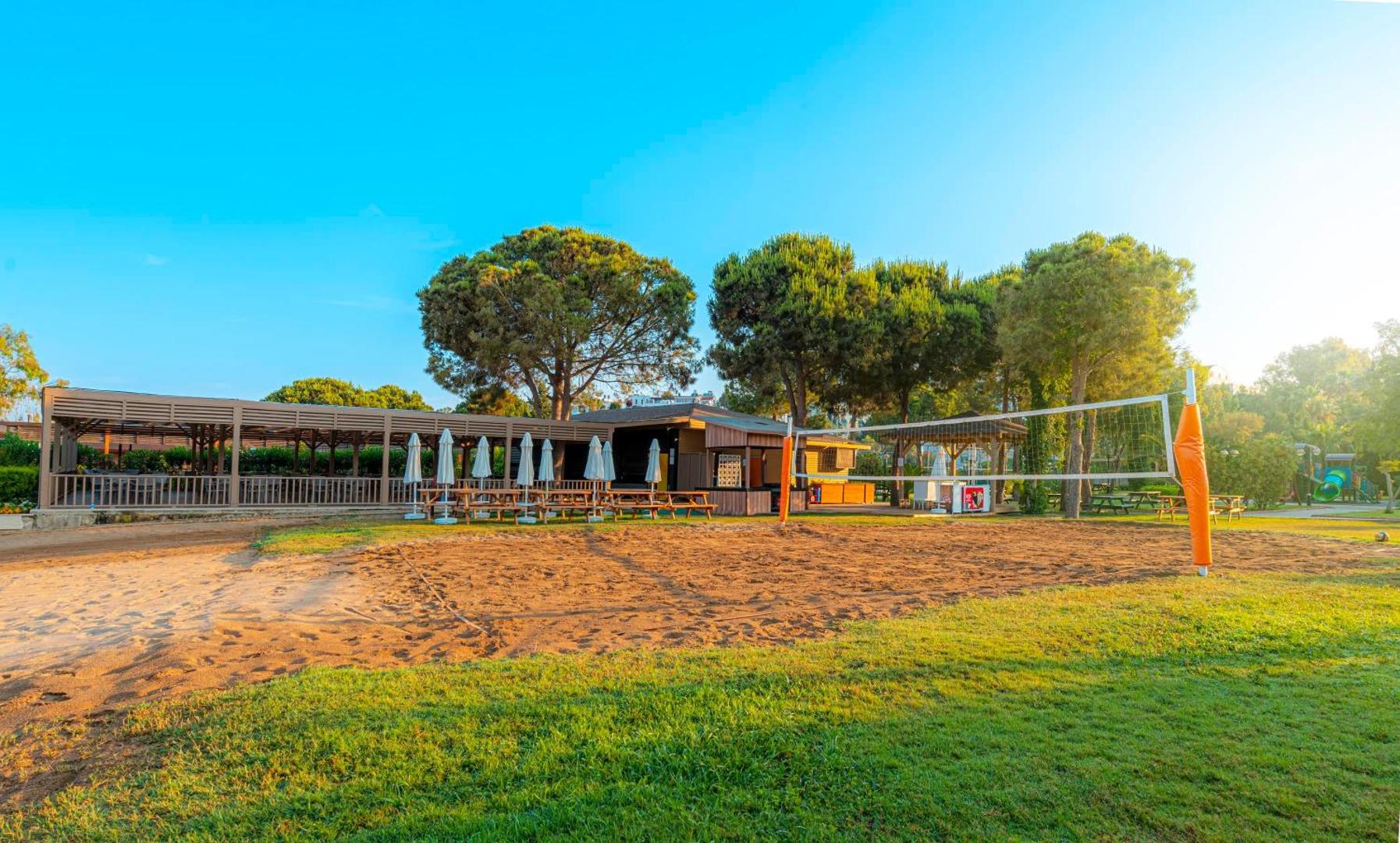 Kirman Sidemarin Beach & Spa pludmales volejbola laukums un āra atpūtas zona ar galdiem un saulessargiem priežu ielenkumā