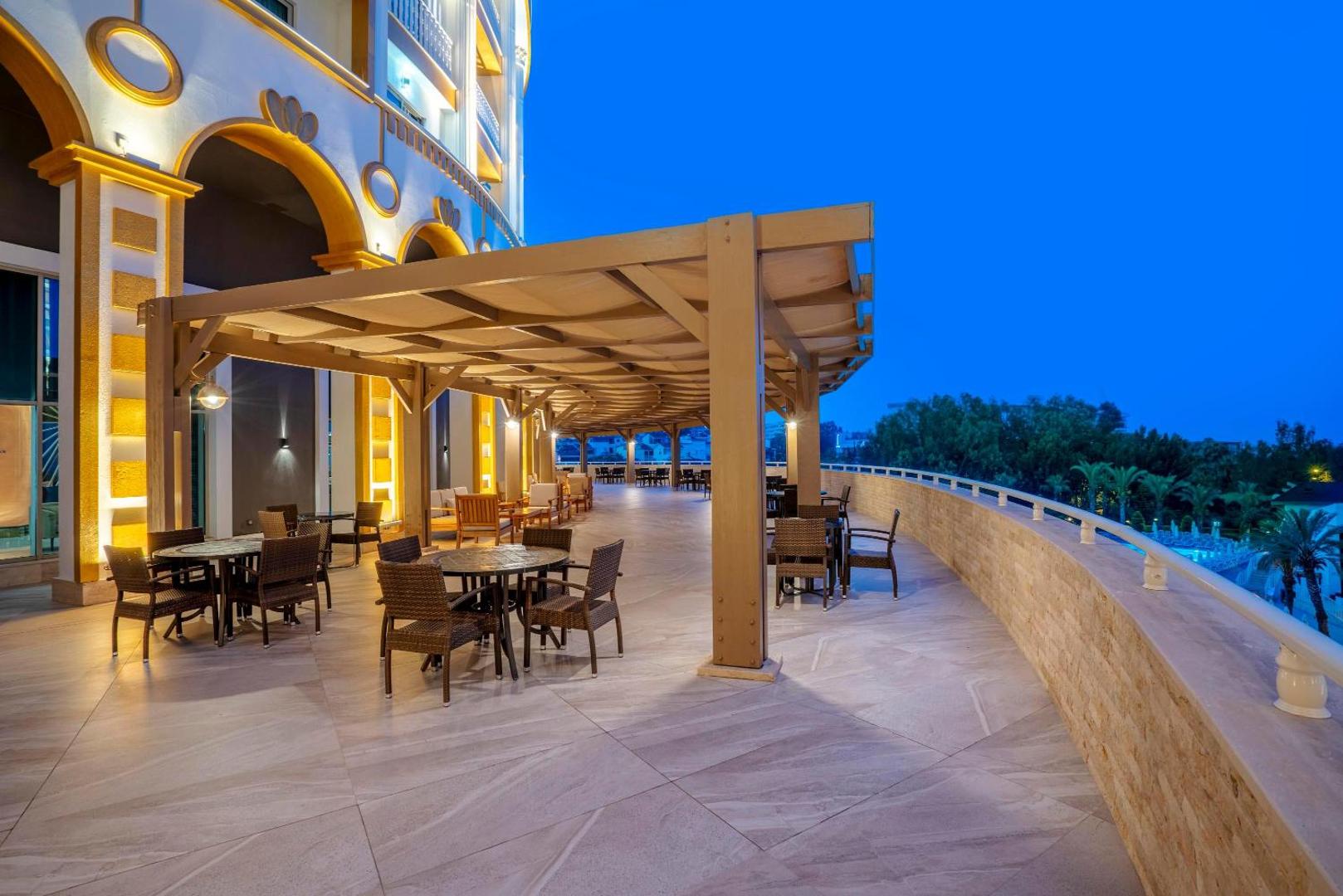 Kirman Sidemarin Beach & Spa viesnīcas āra terase vakarā ar galdiņiem un krēsliem zem nojumes piekrastē, skats uz baseinu
