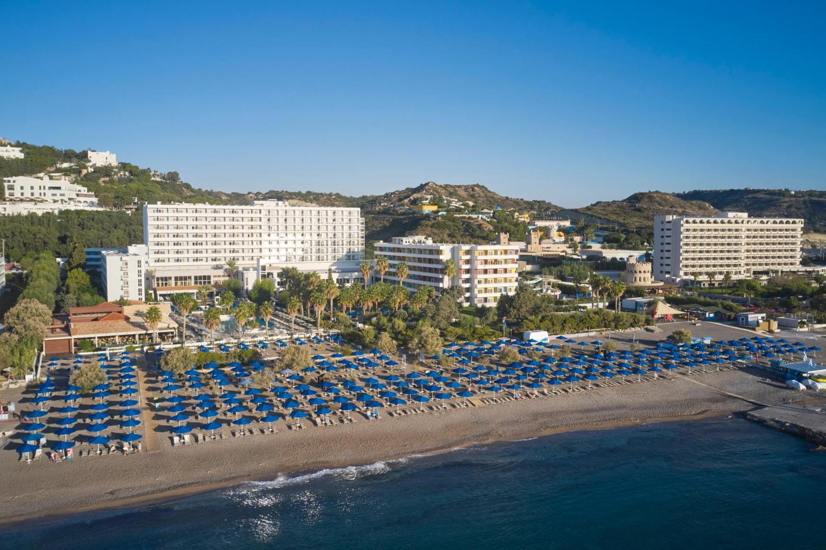 Esperides Beach kūrorta panorāmas skats ar plašu smilšu pludmali, ziliem saulessargiem un viesnīcas korpusiem Rodas salas piekrastē