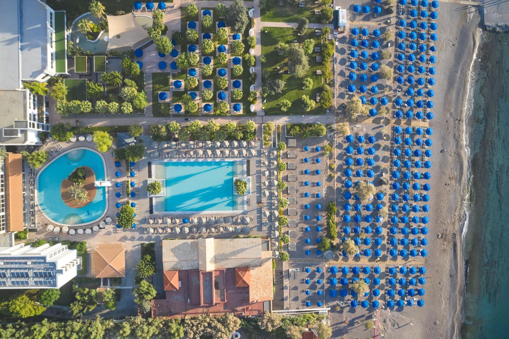 Esperides Beach viesnīcas teritorijas skats no putna lidojuma — baseini, dārzs un plaša pludmales zona ar ziliem saulessargiem