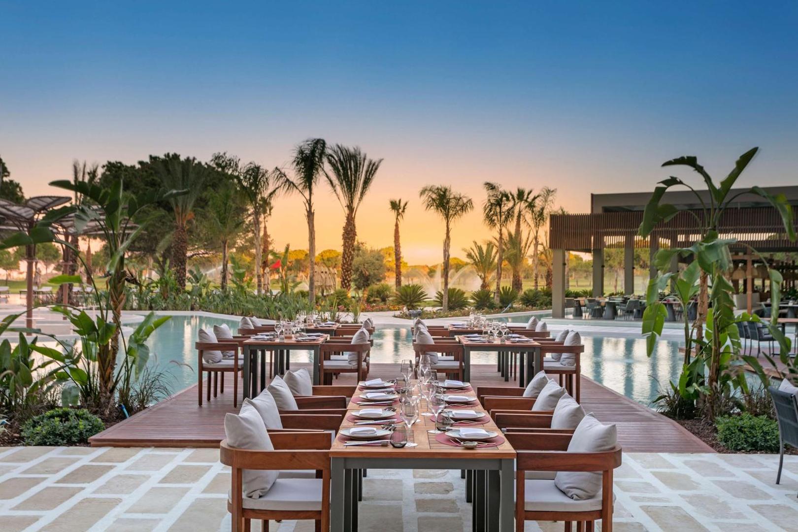 Cullinan Golf & Resort restorāns pie baseina ar koka galdiem, palmām un strūklakām fonā vakara apgaismojumā.