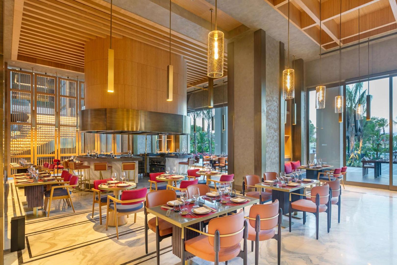Cullinan Golf & Resort moderns iekštelpu restorāns ar atvērto virtuvi, dizaina lampām un elegantiem krāsainiem krēsliem.
