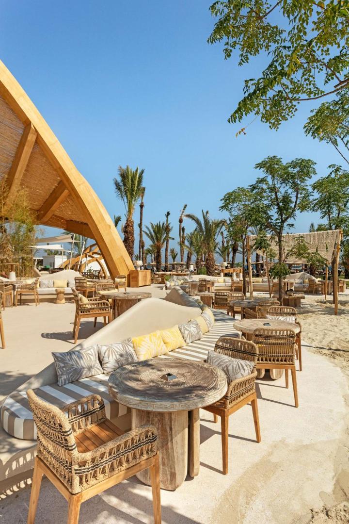 Cullinan Belek Resort pludmales bārs un atpūtas zona ar stilīgām koka mēbelēm, pītiem krēsliem un palmu ēnu.