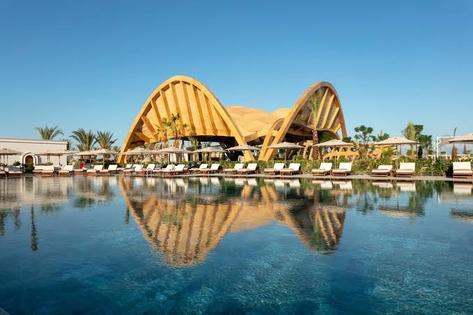 Cullinan Golf & Resort galvenais baseins ar saulessargiem un guļamkrēsliem, fonā redzama moderna koka konstrukcijas ēka un debesis
