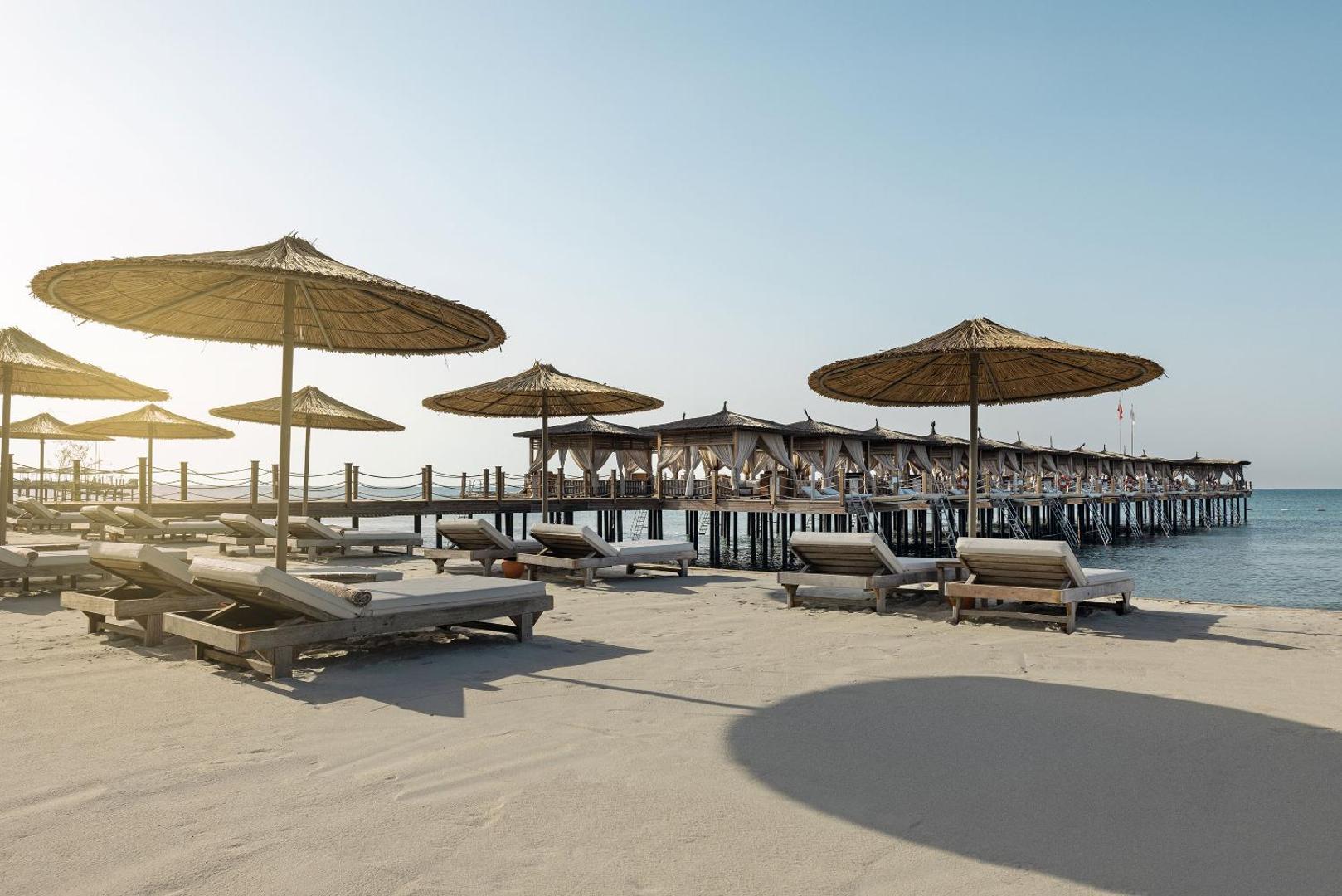 Cullinan Belek Resort privātā pludmale ar koka sauļošanās krēsliem, saulessargiem un garu koka piestātni ar atpūtas namiņiem.