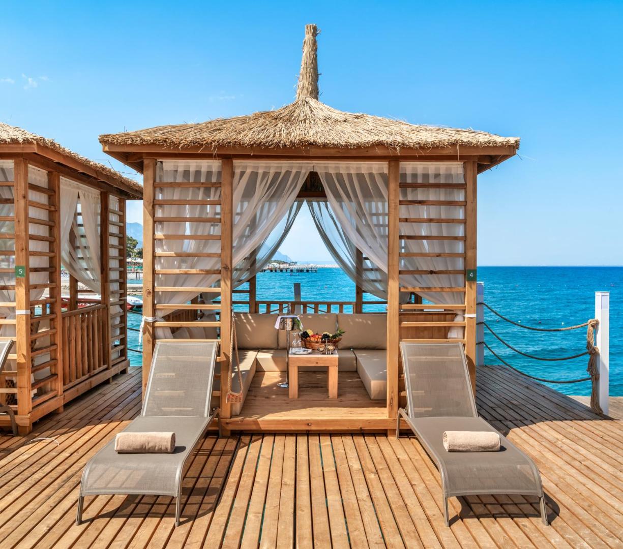 Corendon Playa Kemer privātā koka laipa ar atpūtas bungalo un sauļošanās krēsliem pie jūras — gaiši aizkari un mīksti spilveni