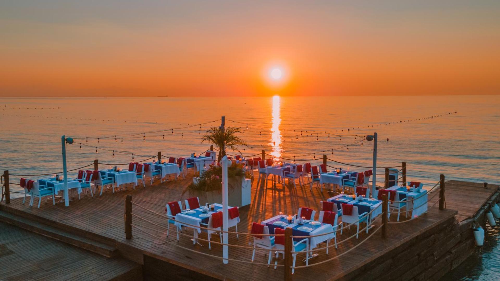 Corendon Playa Kemer a la carte restorāns uz koka piestātnes Vidusjūras krastā saulrieta gaismā ar eleganti servētiem galdiem un sarkanām detaļām.
