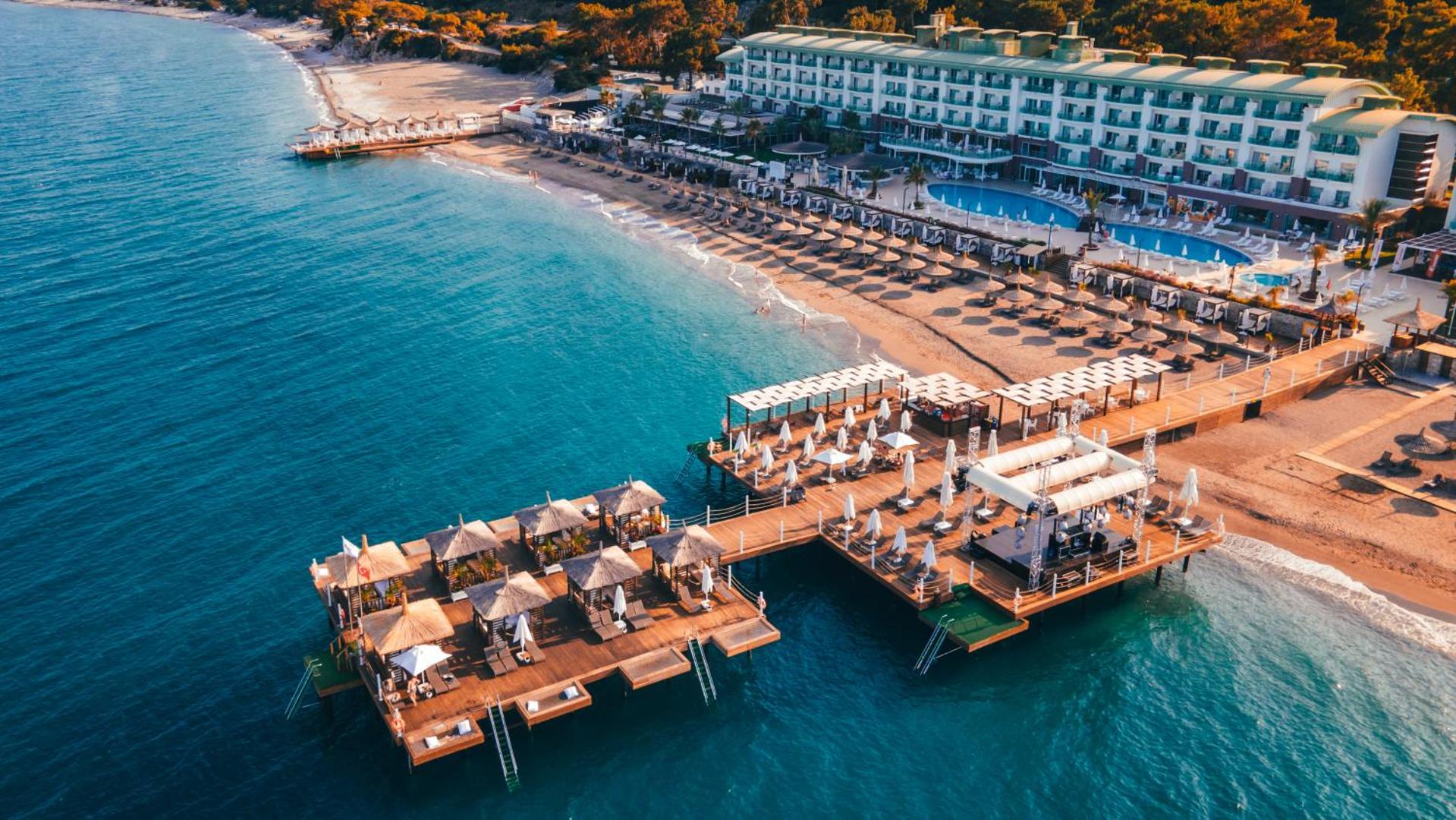 Corendon Playa Kemer skats no putna lidojuma uz viesnīcas ēku, pludmali un koka piestātni ar atpūtas zonām Vidusjūras piekrastē