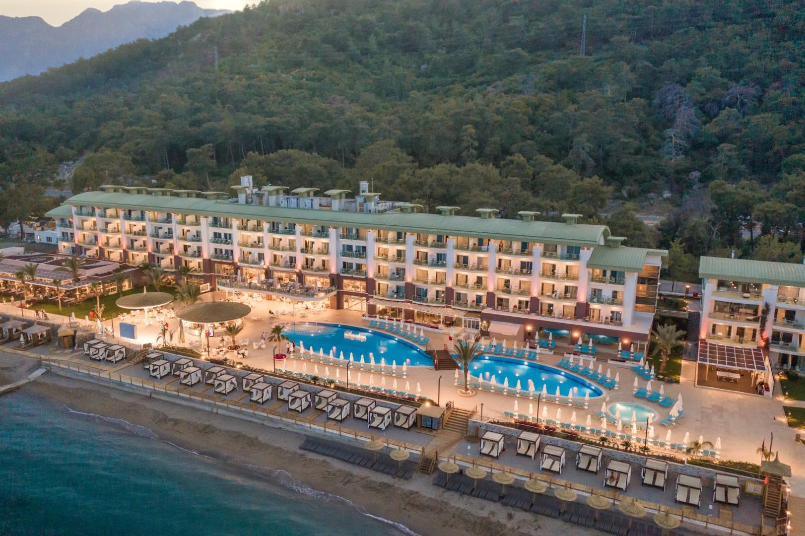 Corendon Playa Kemer viesnīcas fasāde vakarā ar izgaismotiem āra baseiniem, sauļošanās krēsliem un atpūtas zonu piekrastes joslā