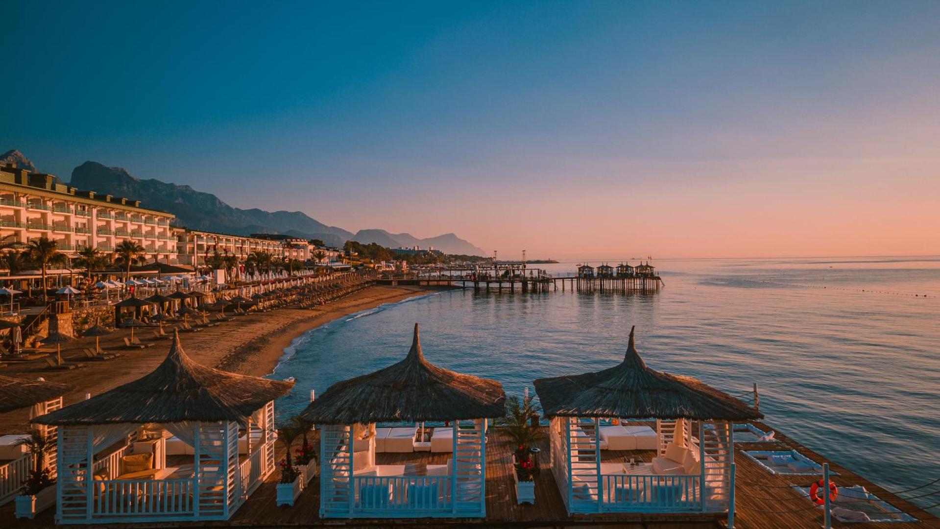Saulrieta skats no Corendon Playa Kemer koka piestātnes uz kalniem un piekrasti ar stilīgām atpūtas teltīm un mīkstiem dīvāniem