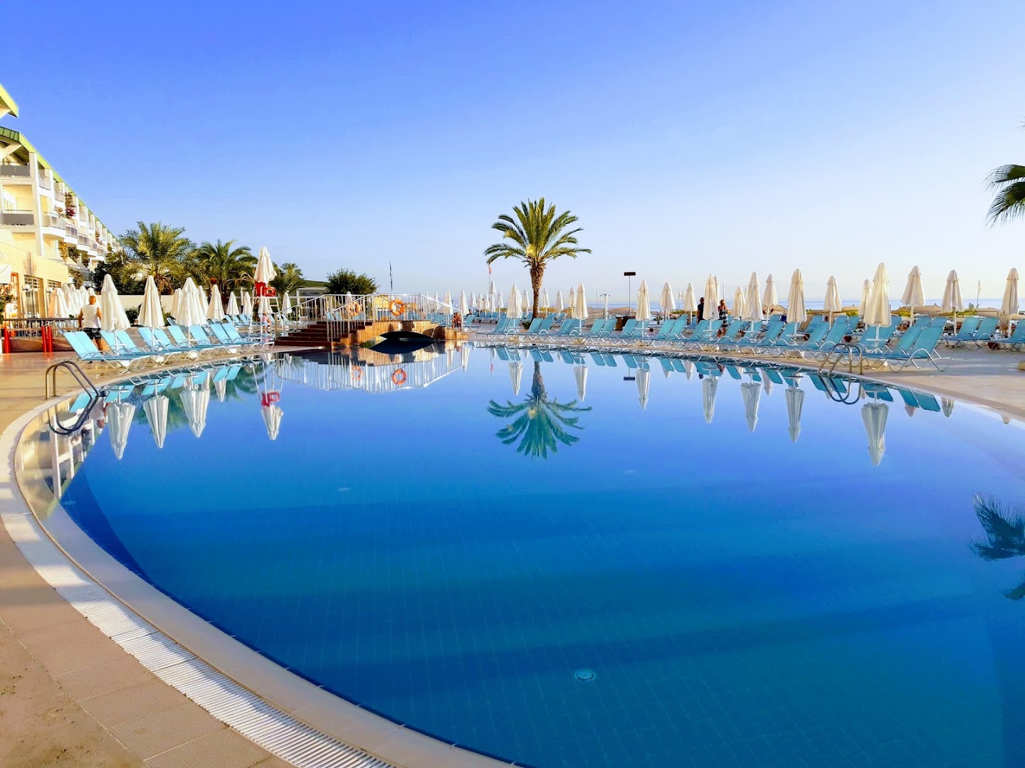 Corendon Playa Kemer lielais āra baseins ar dzidru ūdeni un saulessargiem Kemeras piekrastē — palmu atspulgi ūdens virsmā