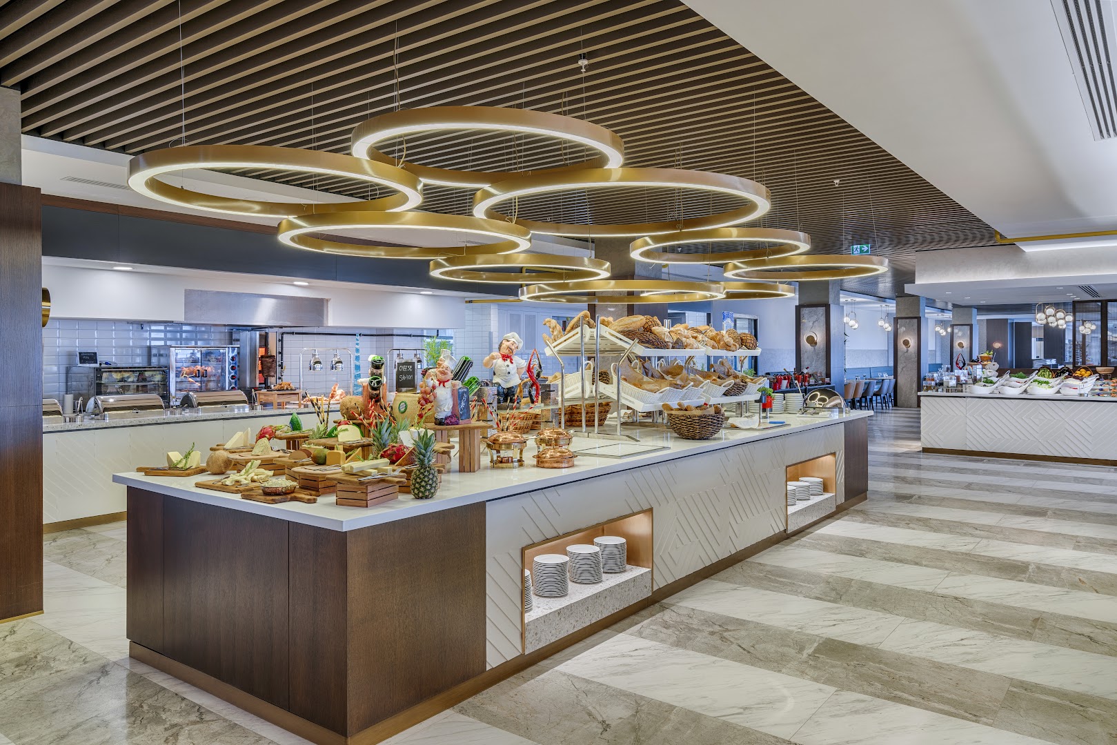 Corendon Playa Kemer galvenā restorāna bufetes zona ar plašu sieru, augļu un svaigas maizes izvēli modernā interjerā ar dizaina gaismekļiem.
