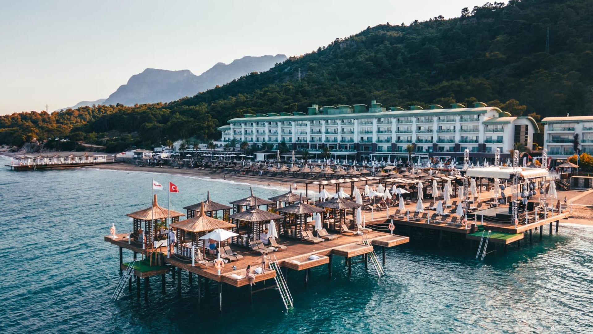 Corendon Playa Kemer viesnīcas ēka un pludmales zona ar koka piestātni, kurā izvietotas atpūtas lapenes un saulessargi pie jūras