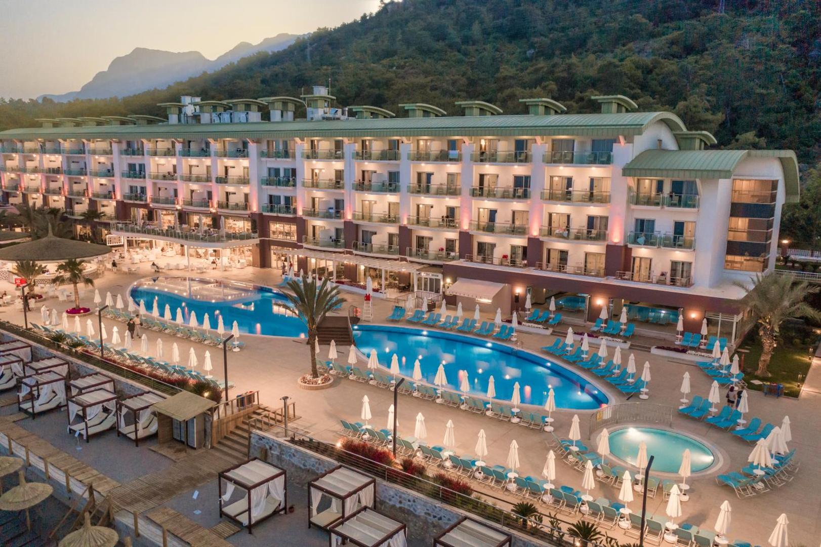 Corendon Playa Kemer viesnīcas ēka un izgaismoti āra baseini vakarā Kemeras reģionā, Turcijā — skats uz kalniem un atpūtas zonu