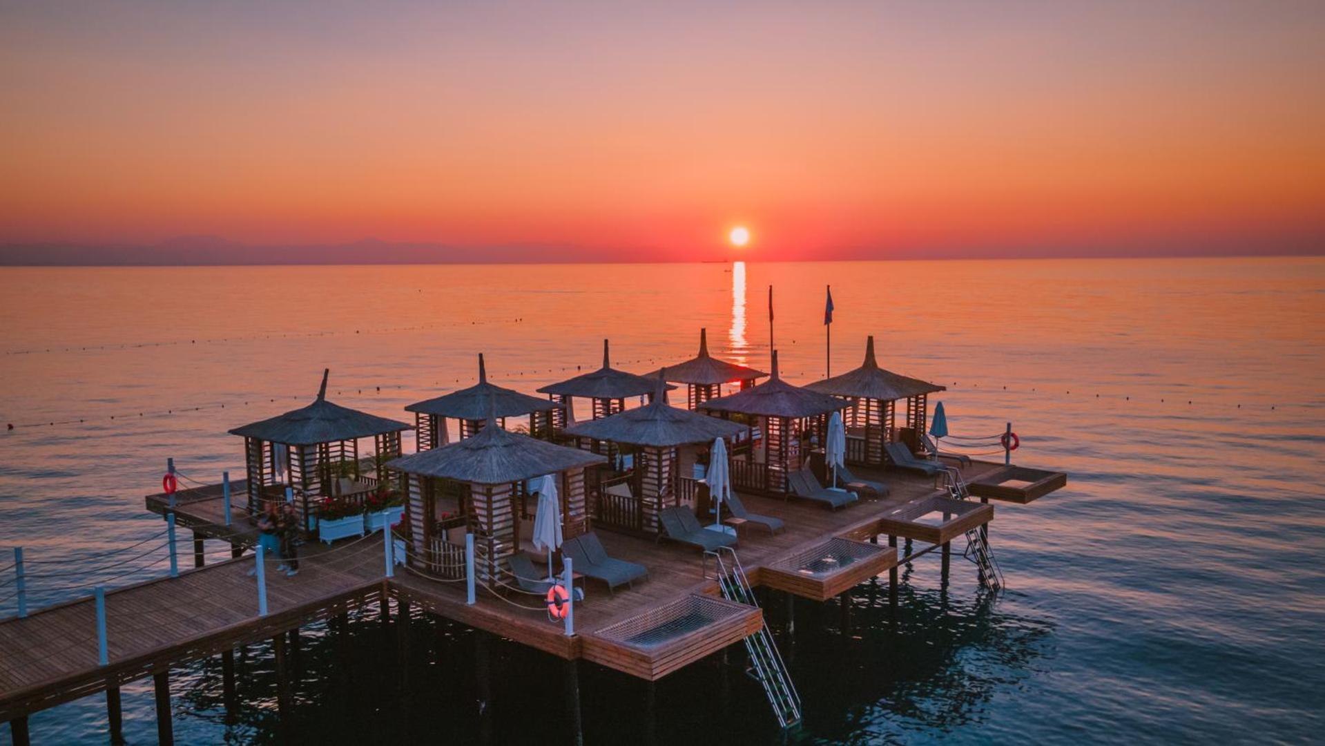 Corendon Playa Kemer koka laipa ar atpūtas namiņiem saulrieta laikā — oranžas debesis virs Vidusjūras piekrastes Kemeras reģionā