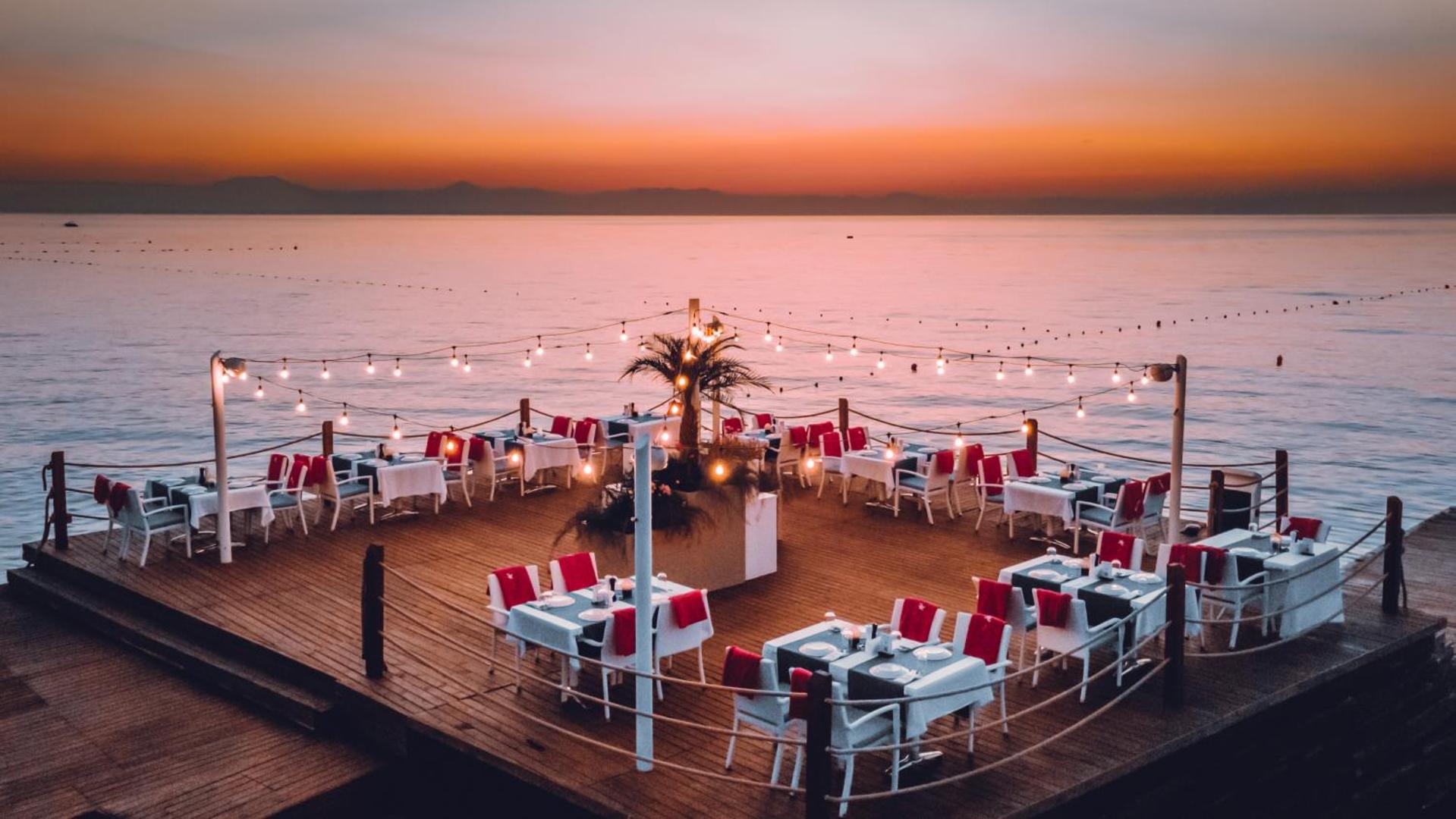 Corendon Playa Kemer restorāns uz koka platformas pie jūras saulrieta laikā, galdi klāti ar baltiem galdautiem un dekorēti ar lampiņu virtenēm.