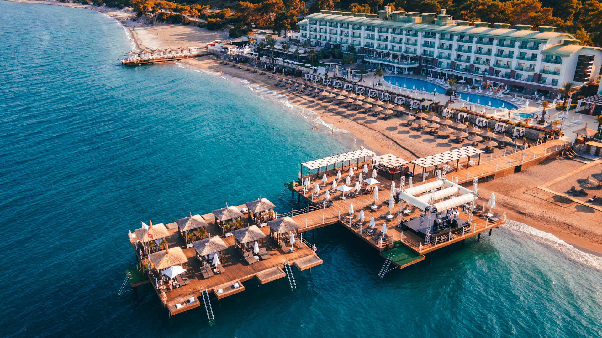 Corendon Playa Kemer panorāmas skats uz viesnīcu, smilšu pludmali un plašo koka piestātni ar bāru un atpūtas vietām