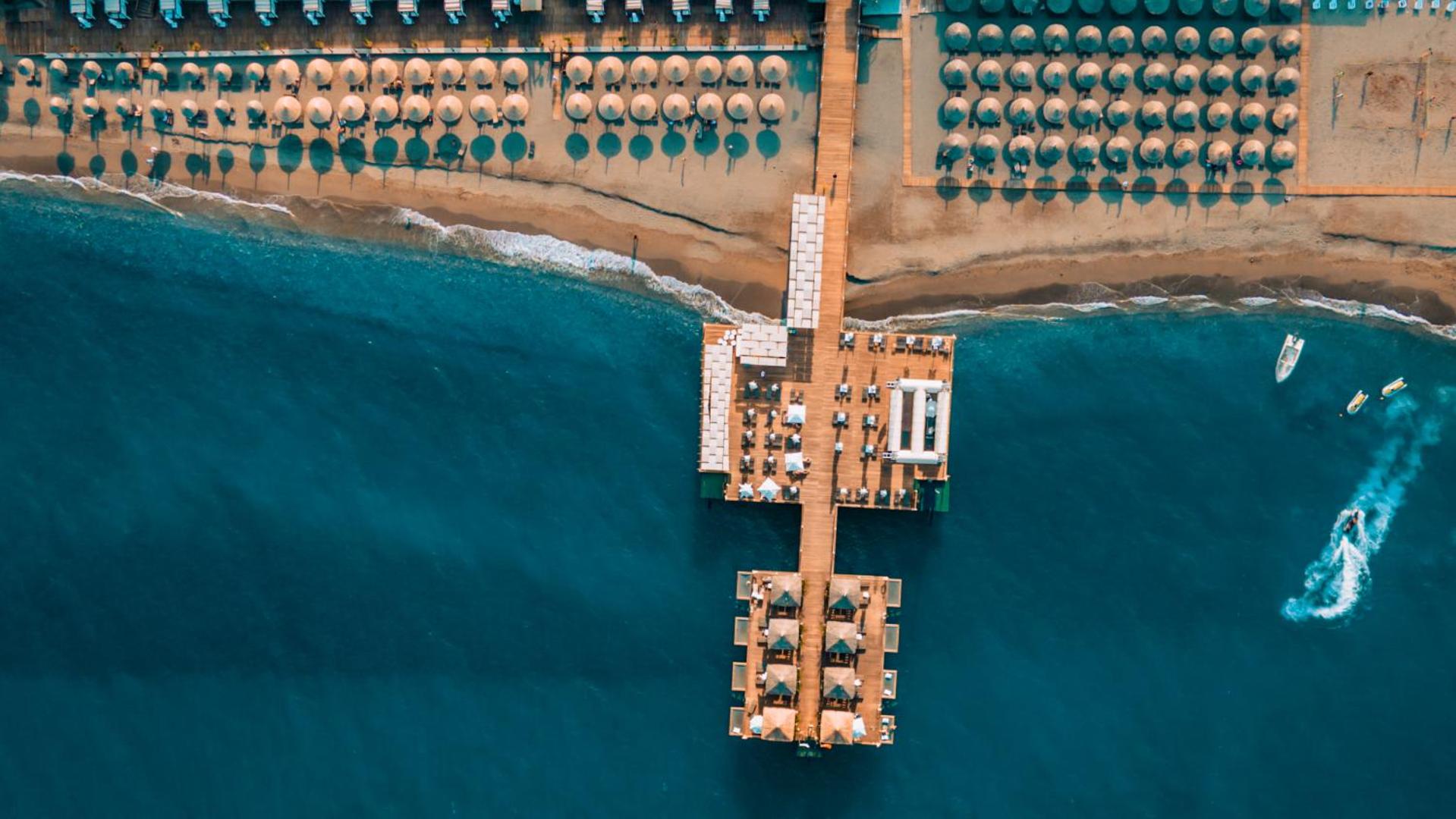 Corendon Playa Kemer viesnīcas pludmales un laipas skats no putna lidojuma — tirkīza ūdens, saulessargi un atpūtas zonas piekrastē